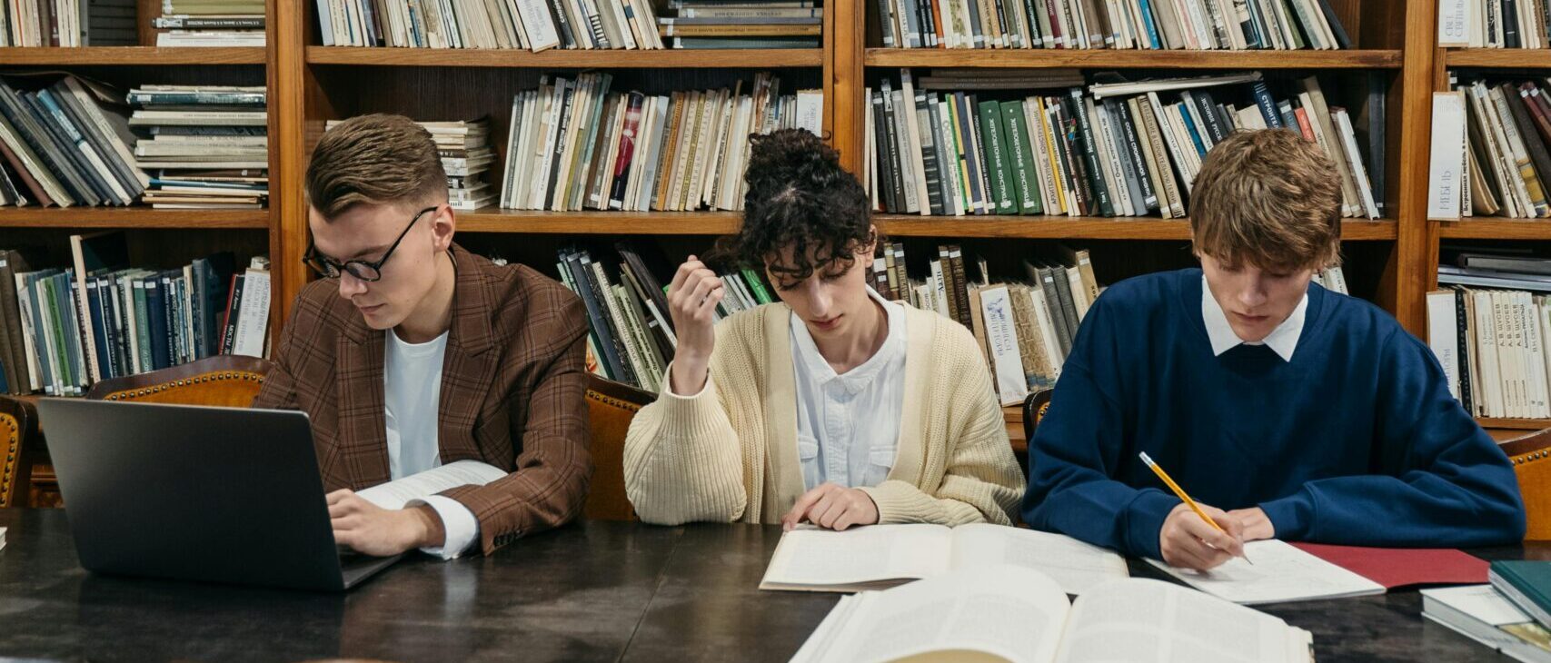 College students engaged in study session at a library table surrounded by shelves of books.